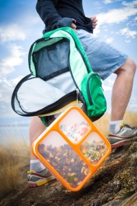 Commercial lifestyle imagery featuring a green hiking backpack and an orange meal prep container in a scenic outdoor setting, shot by Jason Babakaiff.